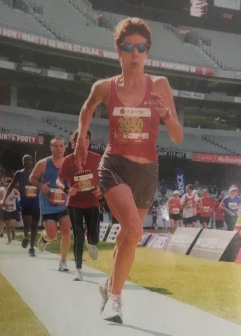 Tina runs across the MCG at the Melbourne Marathon, with the stadium and other runners in the background.
