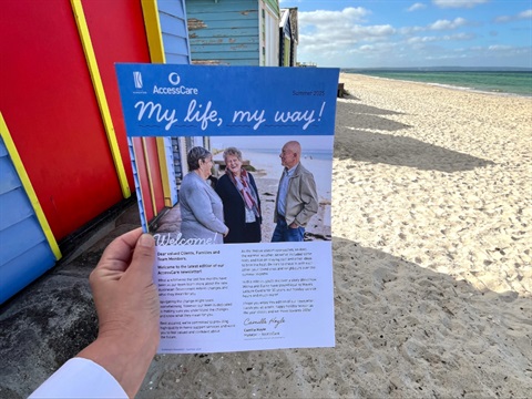 A hand holds a printed newsletter with the beach in the background.
