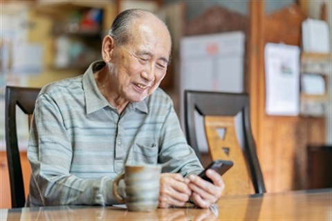 An older man sits at his kitchen table with a cup of coffee, smiling as he uses an app on his smartphone.