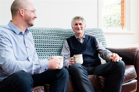 An AccessCare care manager and an older man have coffee and a chat on the couch.