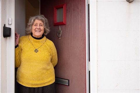 A woman in a bright yellow jumper stands and smile at her front door.