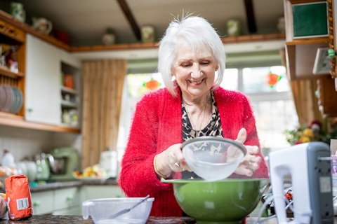 An older woman sifts flour into a bowl in her kitchen,